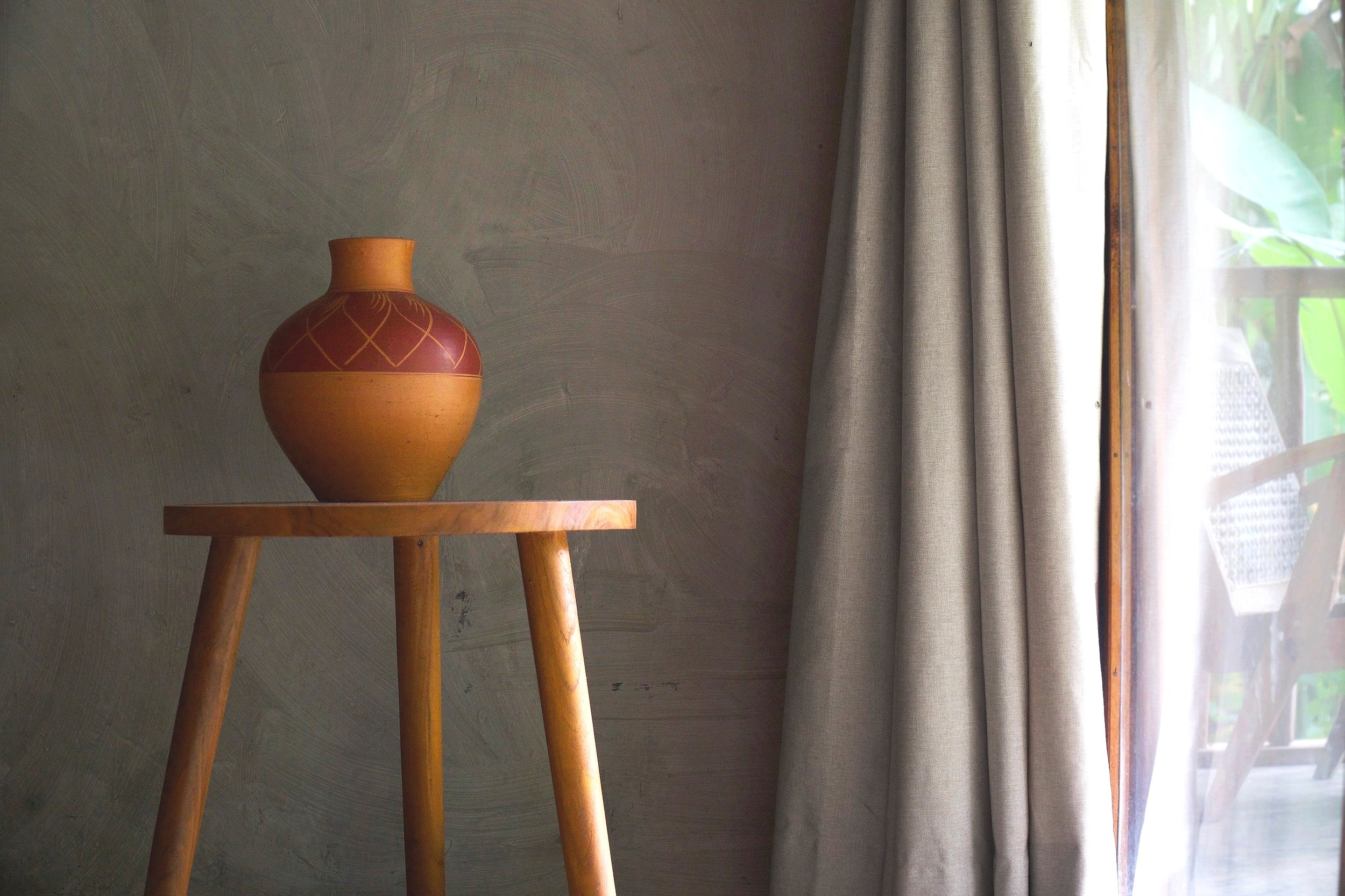 A clay vase on a wooden stool beside a window with curtains, showing the design of the island cottage