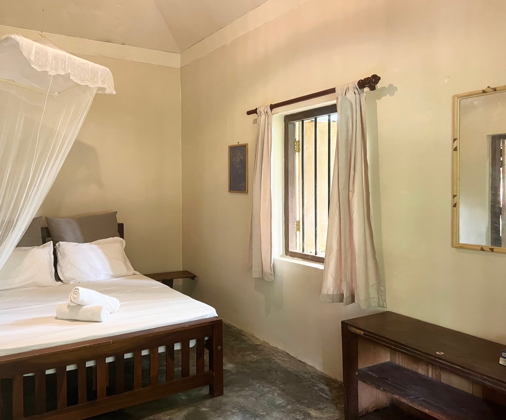 Simple bedroom with a wooden bed, white bedding, mosquito net, white curtains, and a wooden desk.