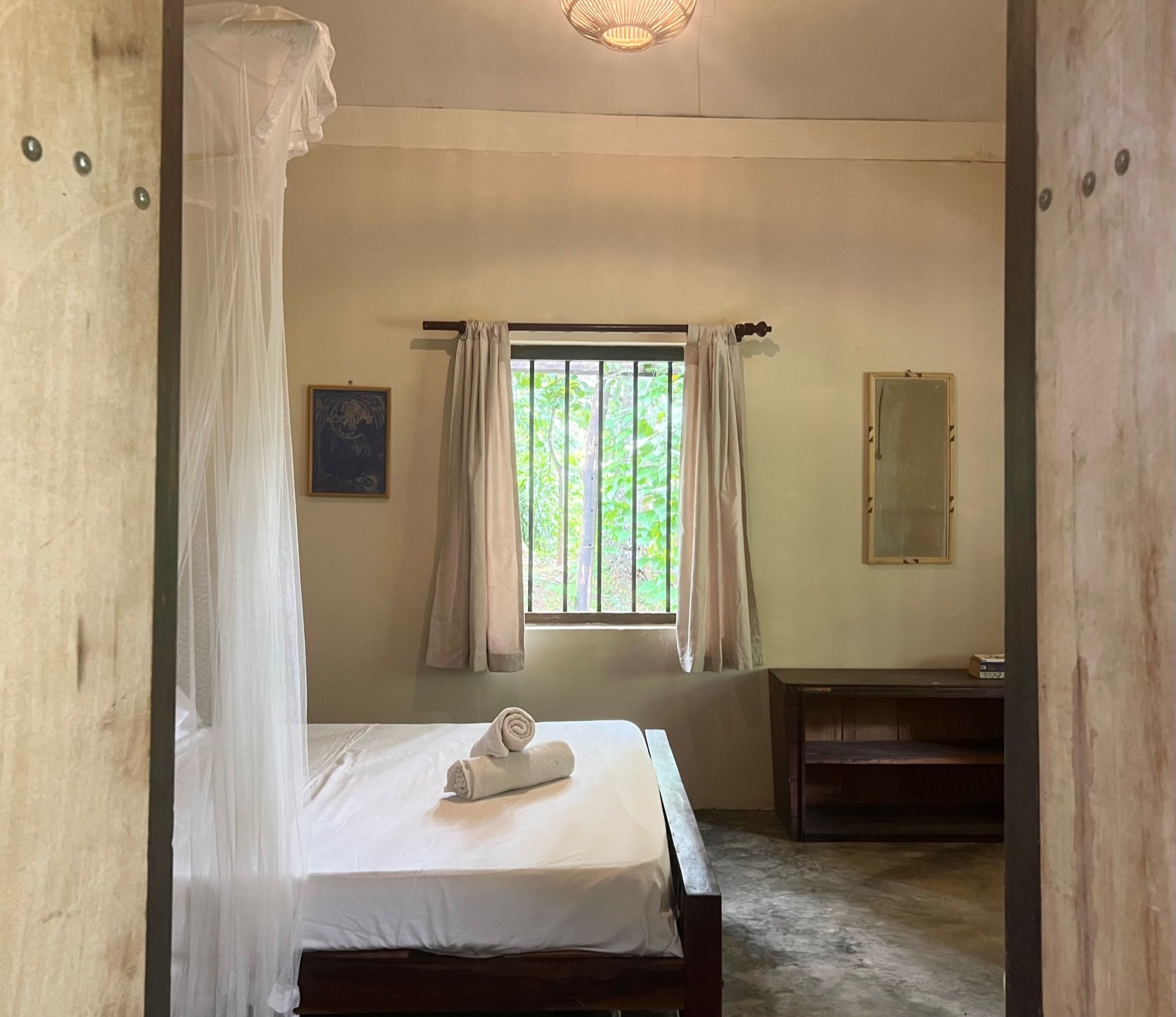 Minimalist bedroom with a double bed, mosquito net, rolled towels, window with curtains, and a small shelf. Room has a rustic, simple design.