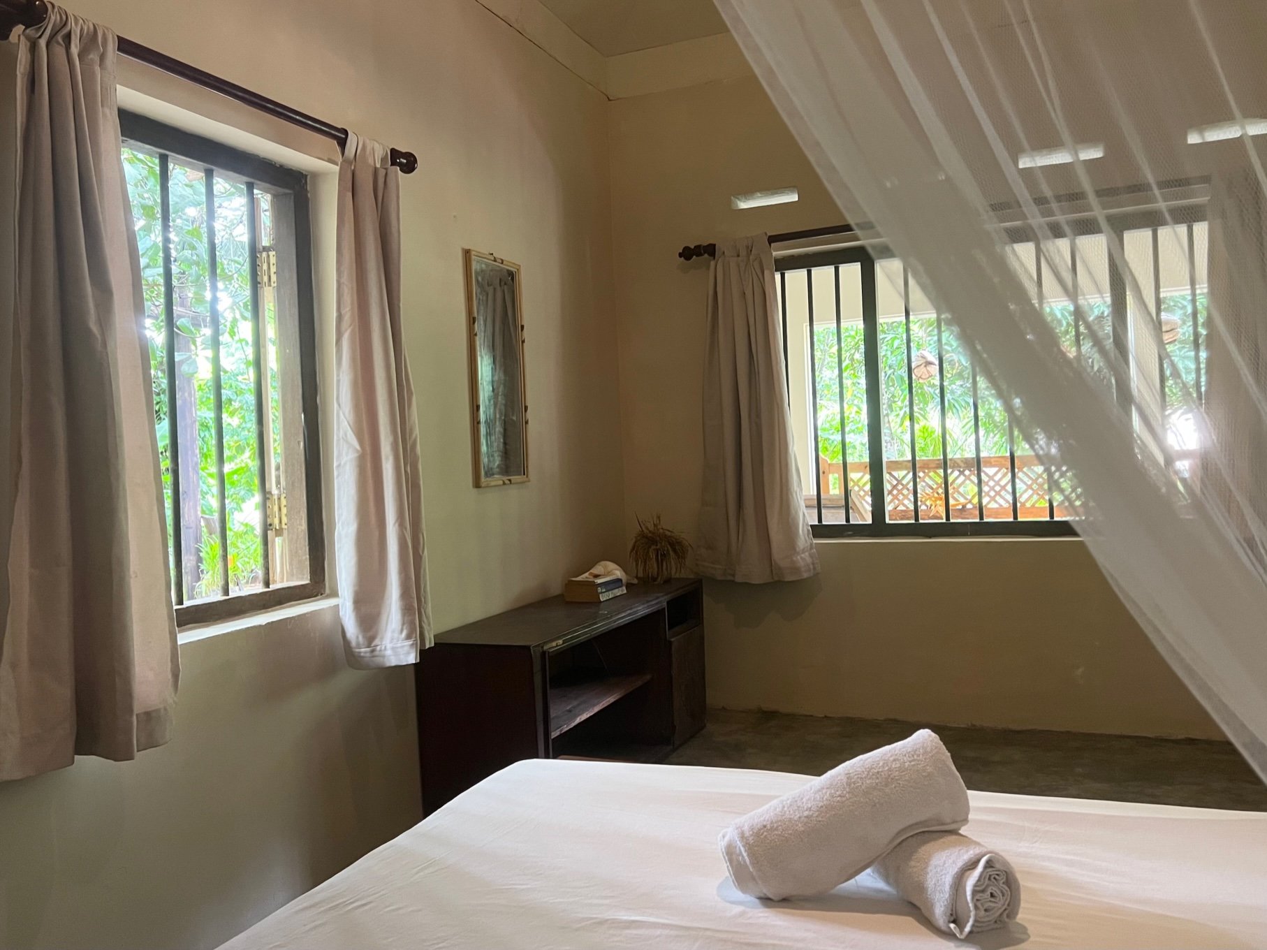 Simple bedroom with a white bed, rolled towels, mosquito net, two windows with curtains, and a mirror on the wall.