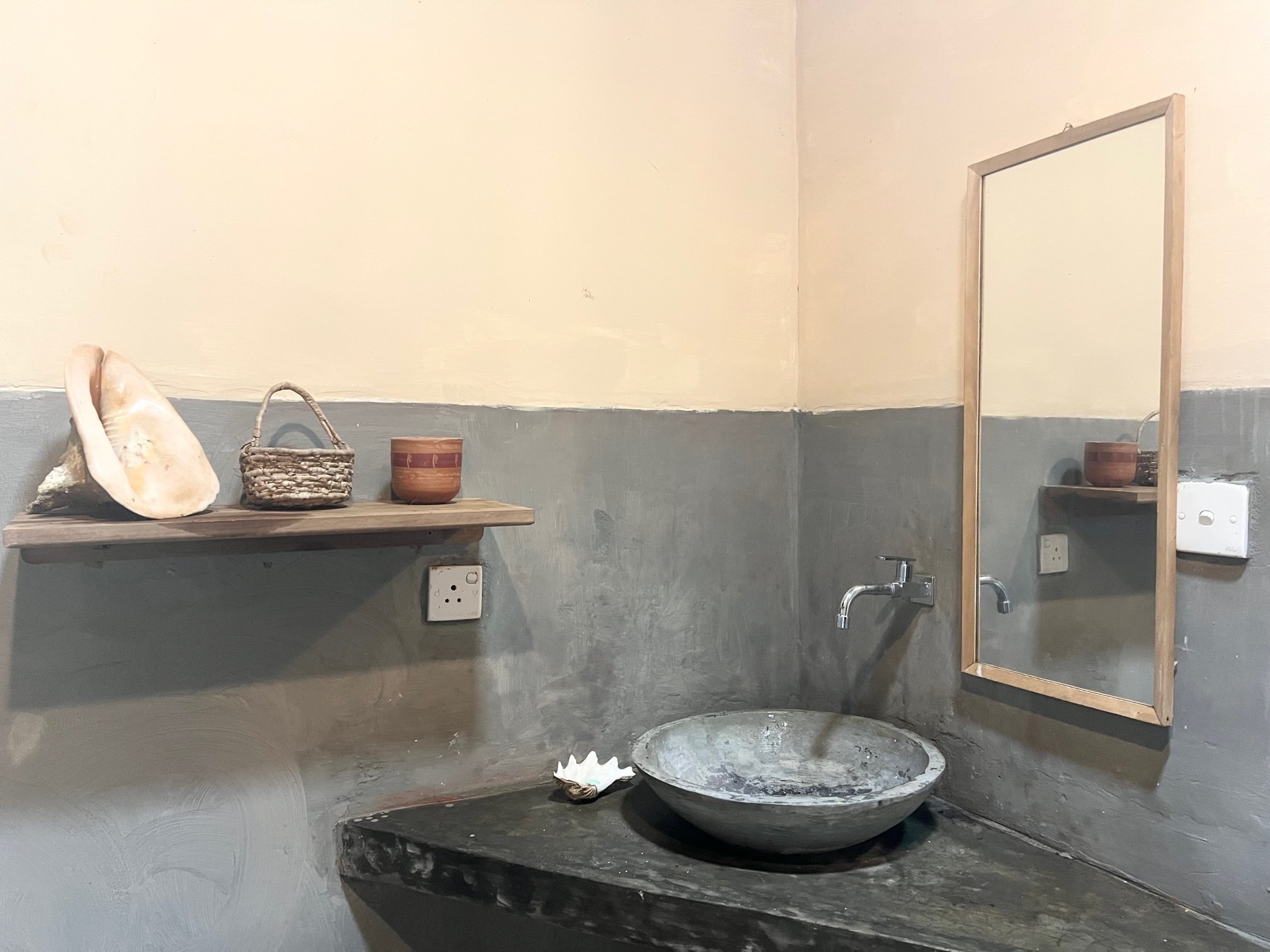 Rustic bathroom with a wooden shelf holding a large seashell, small basket, and a ceramic cup. A concrete sink with a wall-mounted faucet is below a simple wooden-framed mirror. The walls are painted in two-tones: beige upper and gray lower half.