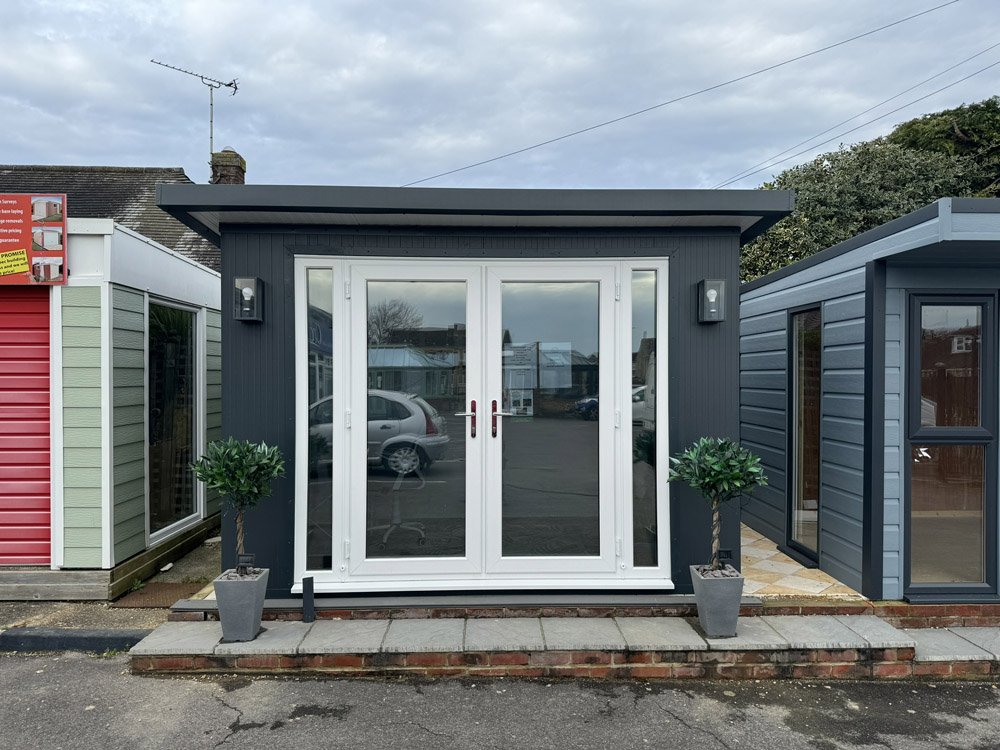 A small modern building with black exterior, glass double doors, and two potted plants outside, set on a paved area in a row of similar buildings.