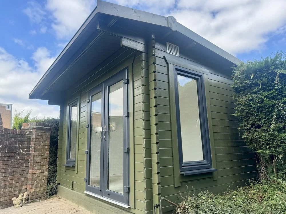 Small green wooden garden shed with glass-paneled door and windows, surrounded by bushes, under a partly cloudy sky.