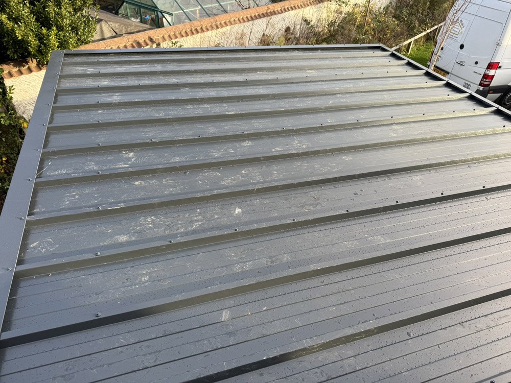 View of a metallic rooftop with vertical ridges, some dirt and debris scattered on its surface, and a garden with plants and a brick border in the background.