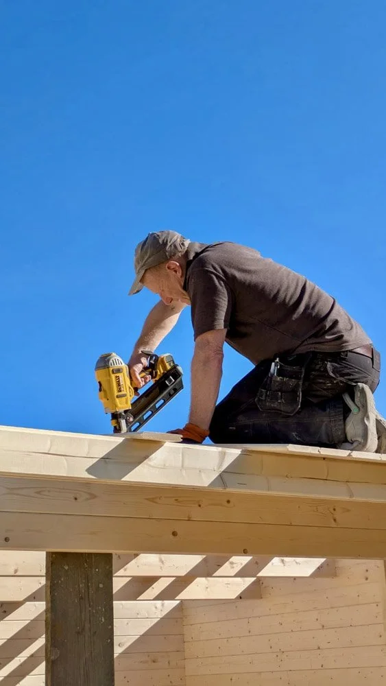 A person kneeling on a wooden platform using a cordless drill against a clear blue sky.