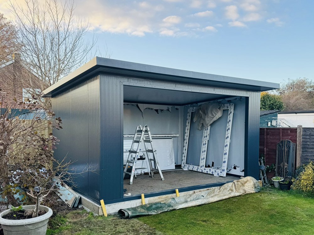 A partially assembled metal backyard shed with a ladder and window frames inside, set in a suburban yard.