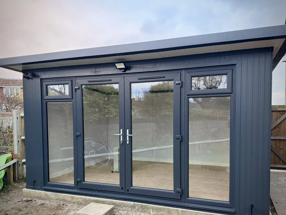 A small dark blue garden shed with large glass doors and windows, situated outdoors on a concrete foundation with a wooden fence and trees in the background.