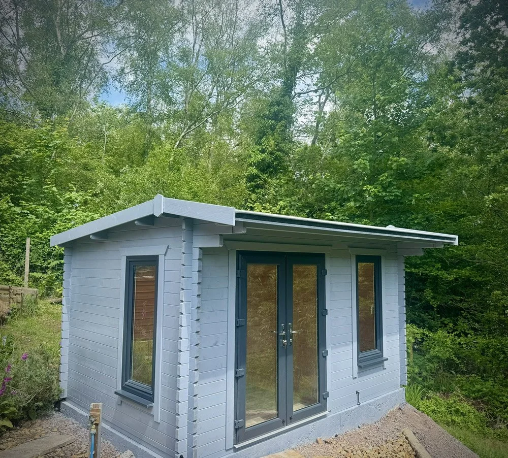 A small, modern shed with gray siding, large glass doors, and two tall narrow windows, situated in a green, wooded area.