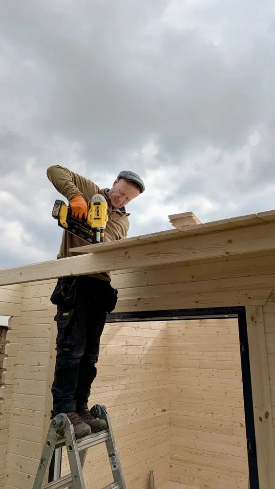 Carpenter working on a wooden structure, standing on a ladder, using a power drill, with cloudy sky in the background.