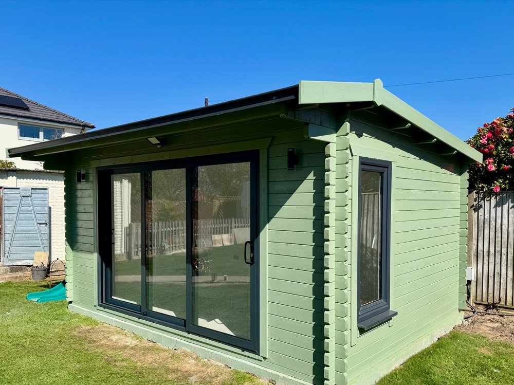 Green wooden garden shed with a sliding glass door and a small window, situated on a lawn with a blue sky background.