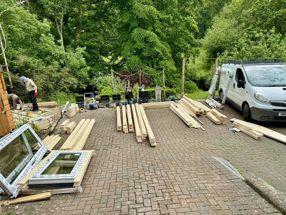 A driveway under construction with wooden planks and tools, a white van parked on the right, and trees and greenery in the background.