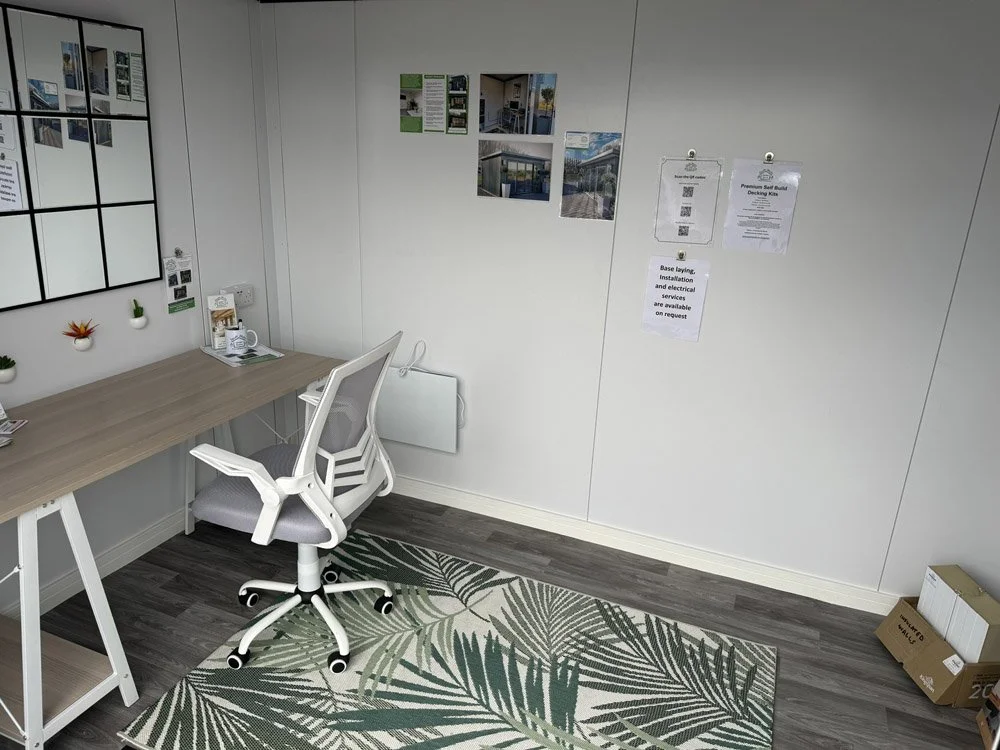 An office or workspace with a desk, a white ergonomic office chair, wall decor including pictures and notices, a patterned rug, and a cardboard box in the corner.