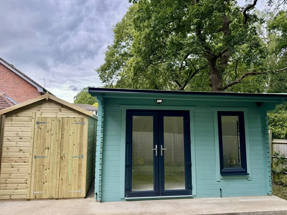 A small light blue wooden building with glass double doors and a window, situated next to a light wood shed in a backyard with trees and a fence.