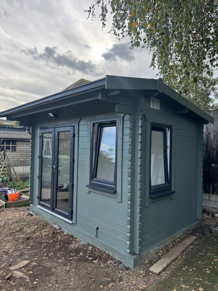 A small, newly built green wooden garden house with black-framed windows and a double door, situated on a dirt patch in a backyard, with trees and a cloudy sky in the background.