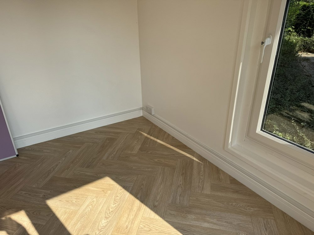 A corner of a room with hardwood herringbone flooring, a white wall, a large window, and a view of greenery outside.