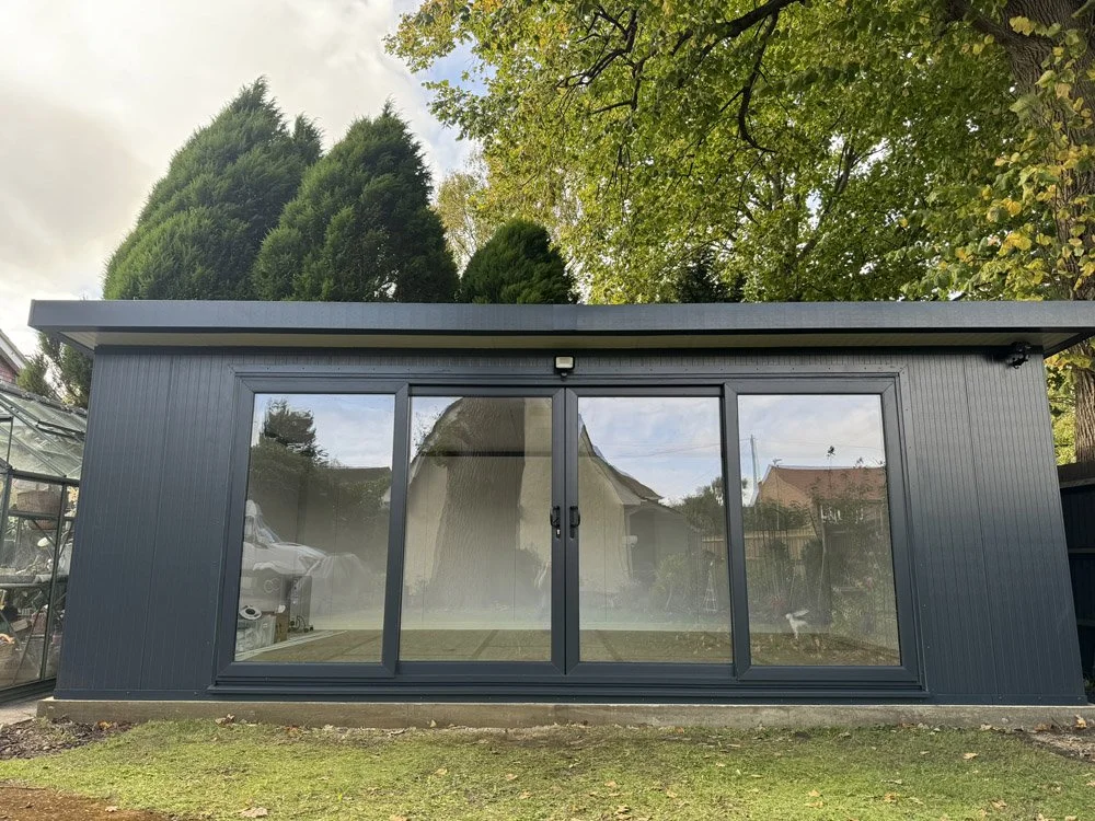 A small, modern, dark gray shed with large glass sliding doors, situated on a grassy yard with trees in the background.