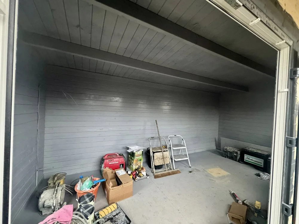 Empty garage with various tools and boxes on the floor, interior with gray walls and ceiling, open garage door.