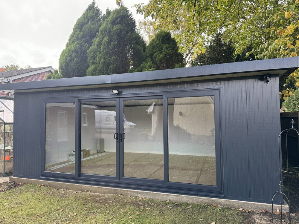 A modern outdoor garden shed with dark grey metal siding and large glass sliding doors, situated in a backyard with grass, trees, and neighboring houses.
