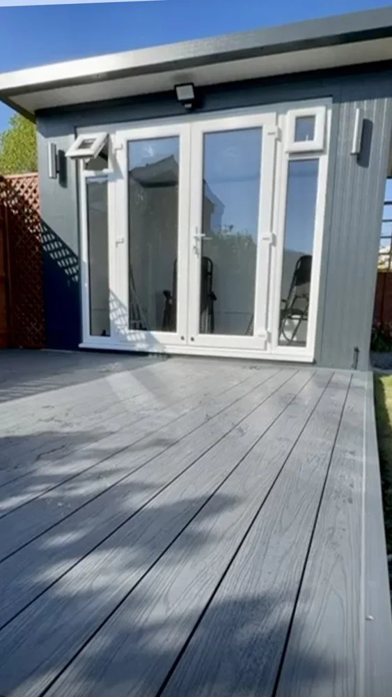 Wooden deck with sliding glass door leading into a house, with outdoor chairs visible inside, next to a gray exterior wall and a small window.