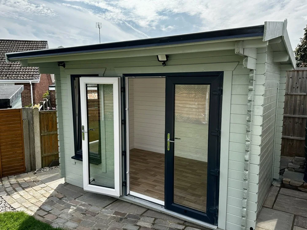 Small backyard shed with double glass doors, one door open, overlooking a brick-paved patio, fenced yard, and neighboring houses under partly cloudy sky.