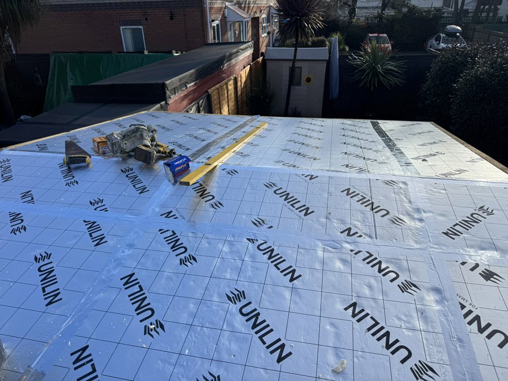 A construction site on a rooftop showing insulation material labeled 'Unilin', along with tools and a level, with neighboring houses, trees, and parked cars visible in the background.