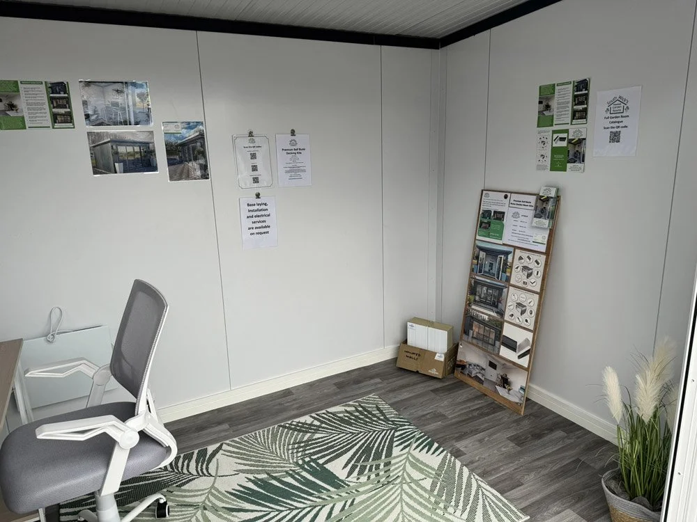 Office corner with a gray swivel chair, a leaf-patterned rug, informational brochures, and posters on the wall about construction or renovation projects, with some boxes and a wooden display stand in the corner.