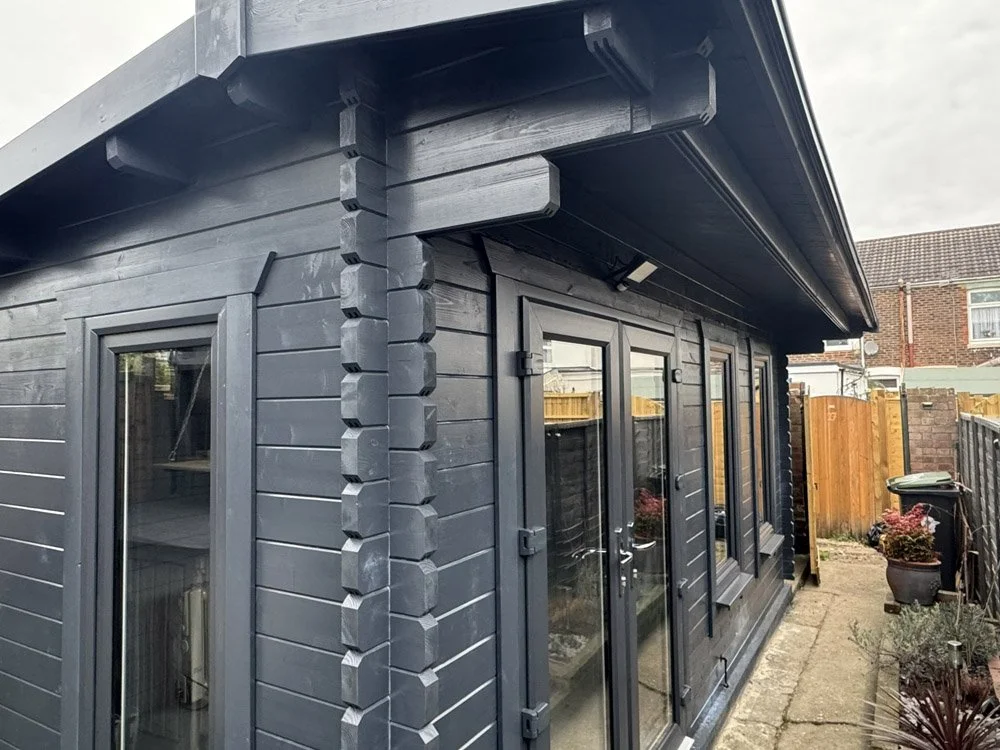 A black wooden garden shed with double glass doors, a window, and a dark roof, situated in a backyard with a small concrete path, potted plants, and a wooden fence.