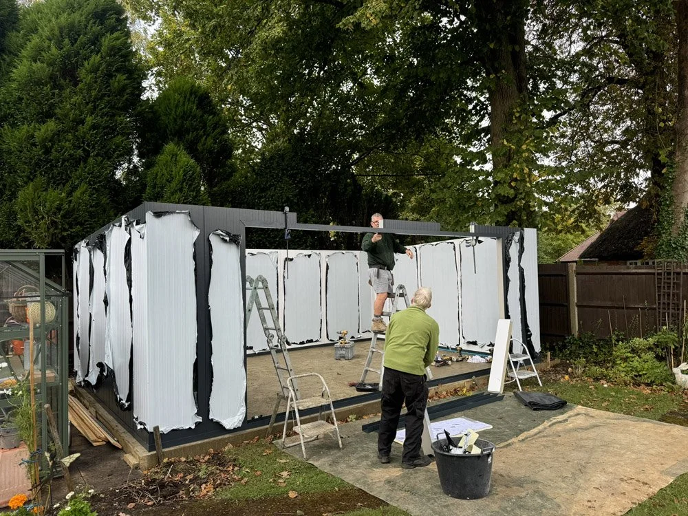 Two men installing a black and white outdoor shed or structure, with one on a ladder and the other standing on the ground, surrounded by tools and construction materials.