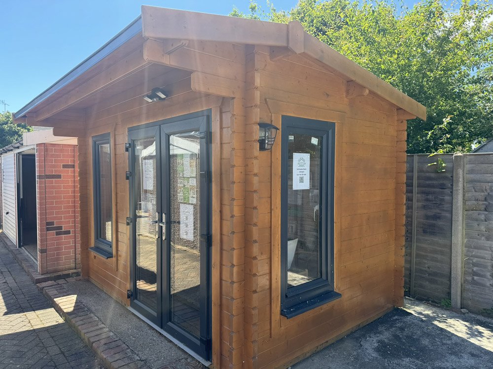 Small wooden garden shed with glass double doors and a window on side, located outdoors with a wooden fence and trees in background.
