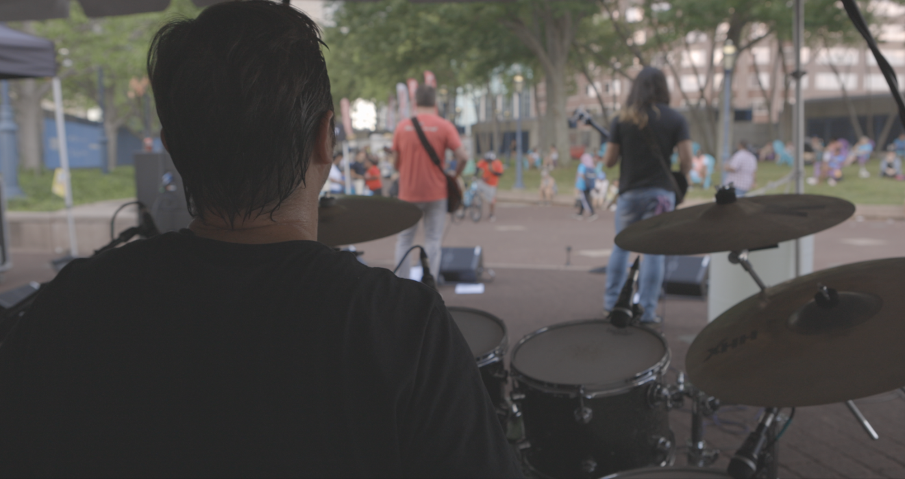 A musician playing drums on a stage in an outdoor park while people walk in the background.