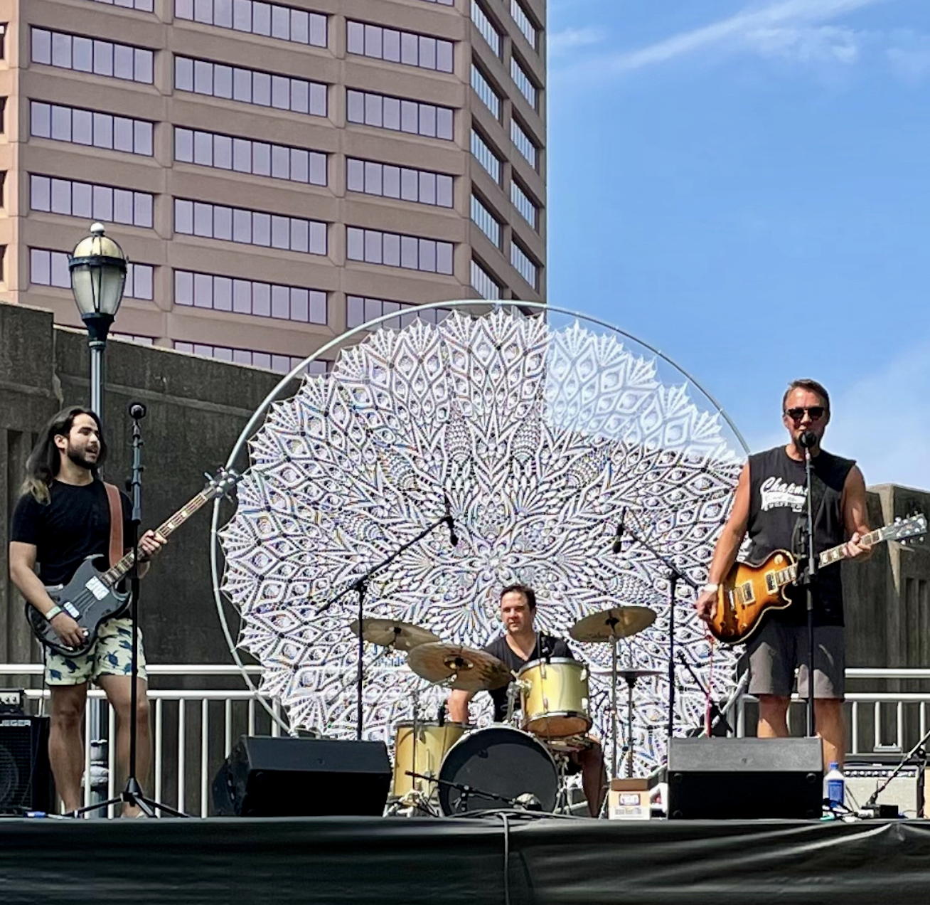 A band performing on an outdoor stage with three members playing instruments. The drummer is centered behind a decorative circular backdrop, with one guitarist to the left and another to the right, under a clear blue sky.