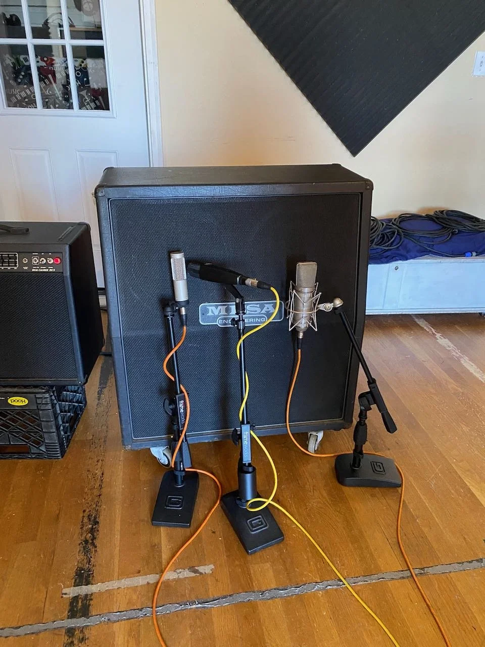 Three microphones on stands positioned in front of a large black speaker, connected by orange and yellow cables, inside a room.