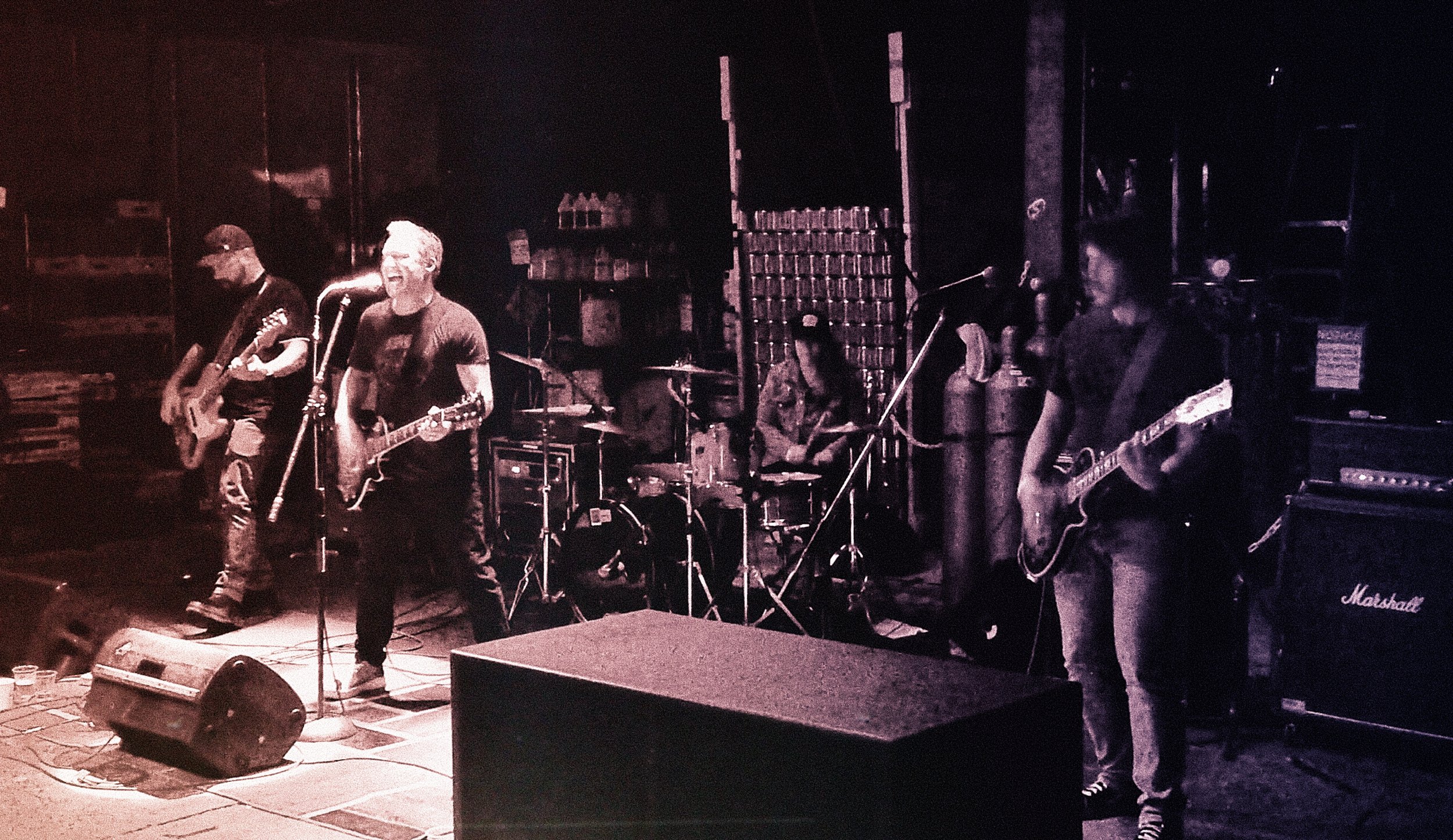 A band performing live on stage with members playing guitars and drums, with music amplifiers and equipment in the background.