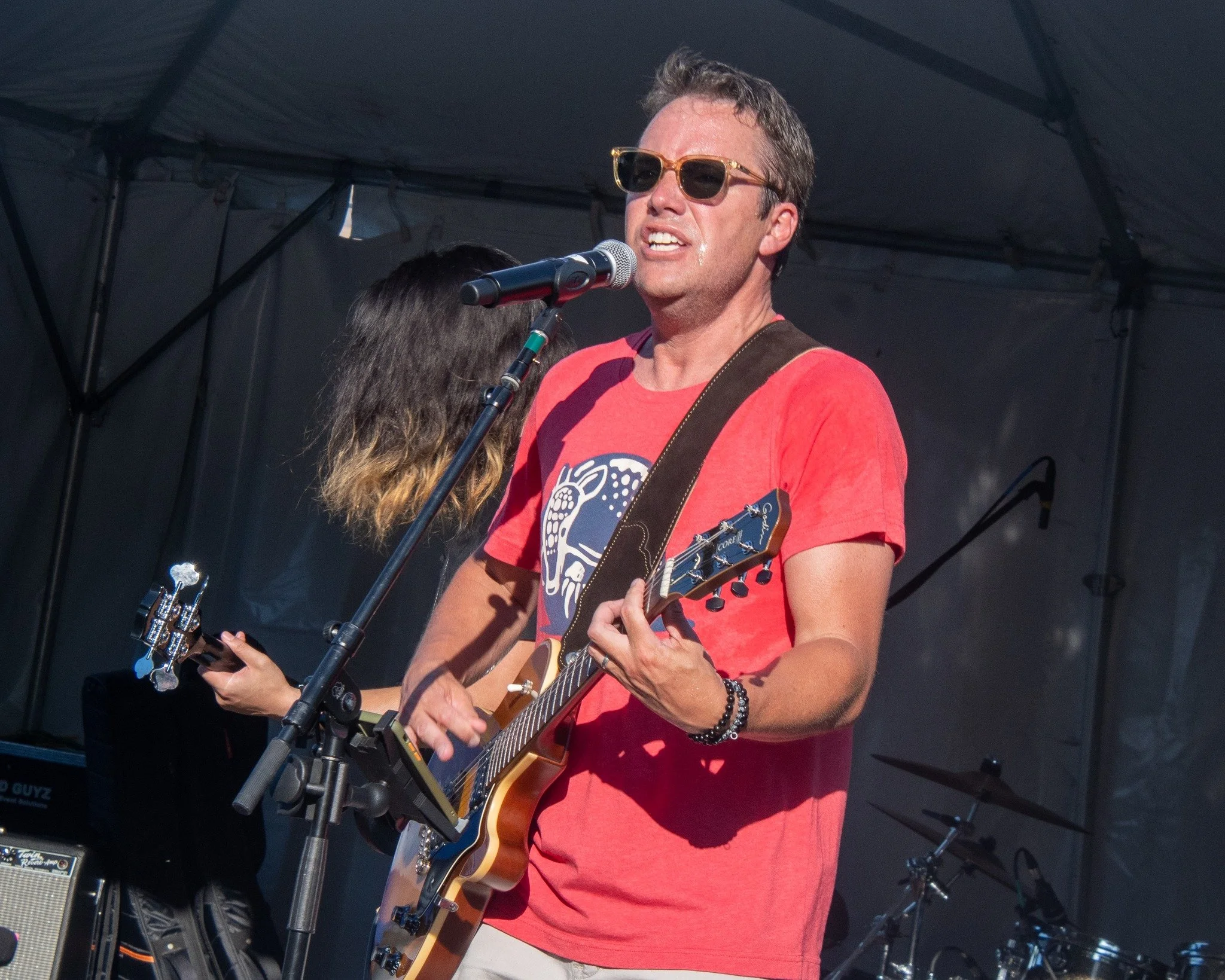 A man wearing sunglasses and a red t-shirt is playing an electric guitar and singing into a microphone at an outdoor concert.