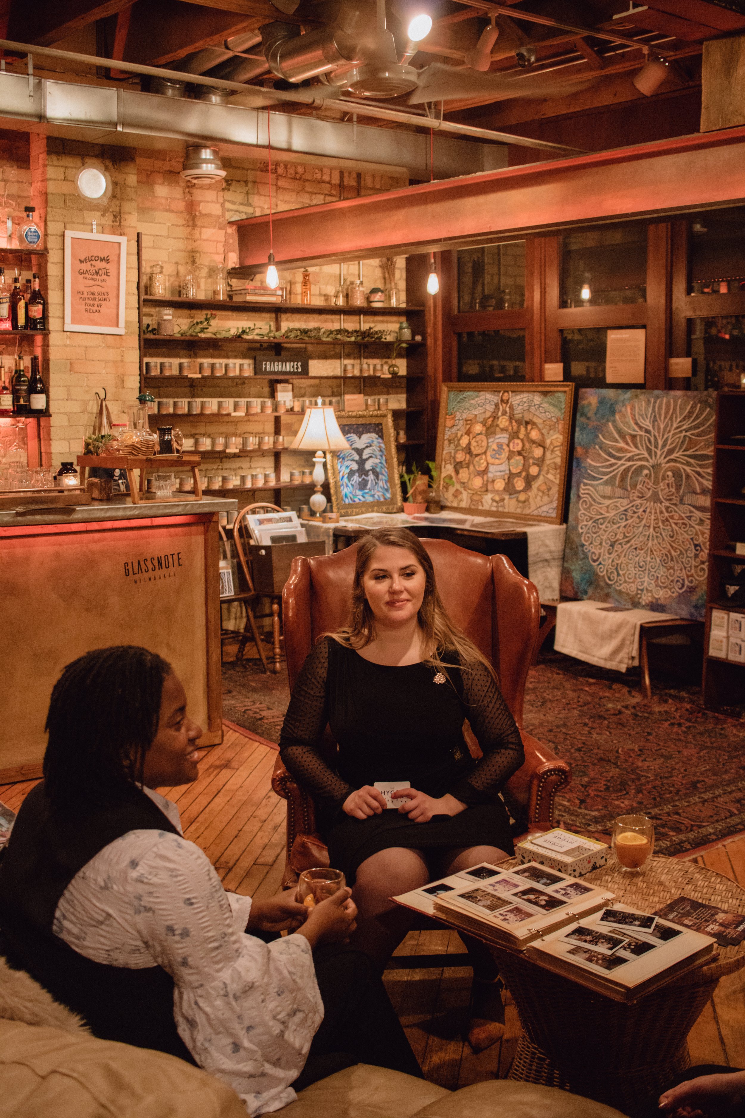  Milwaukee artist and curator, Lauren Marie Nitka, sitting in an arm chair with the art of  Jonathan Shaw of Yehonatan Art  in the background, meets with VIP guest at Hygge: A Pop Up Gallery at  Glassnote Candle Bar,  in Walker’s Point, WI in a cream