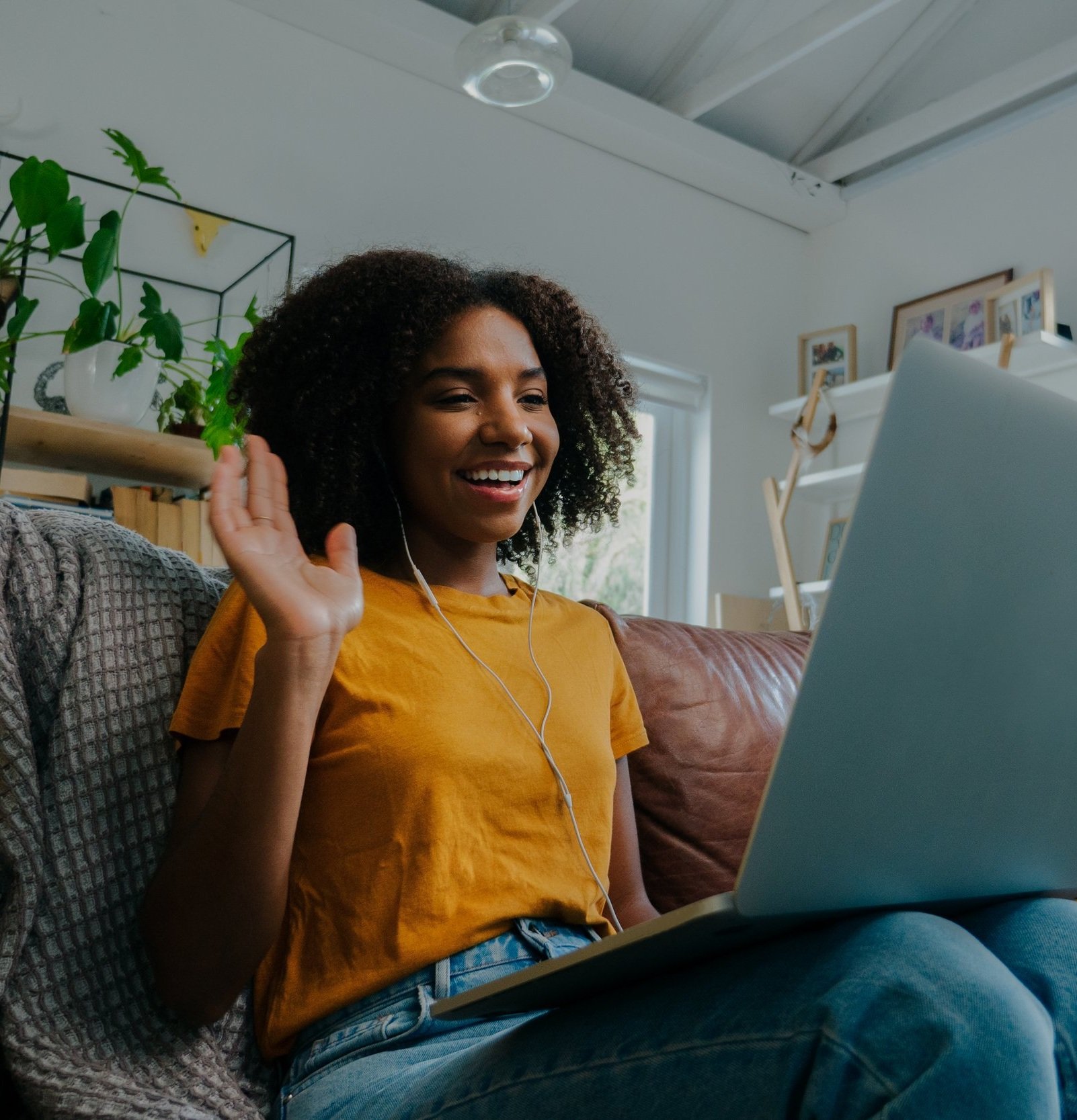 Female-student-wearing-earphones-on-video-call-waving-at-class-mates-through-laptop-sitting-on-leather-couch-in-lounge-1271320826_4608x3072.jpg