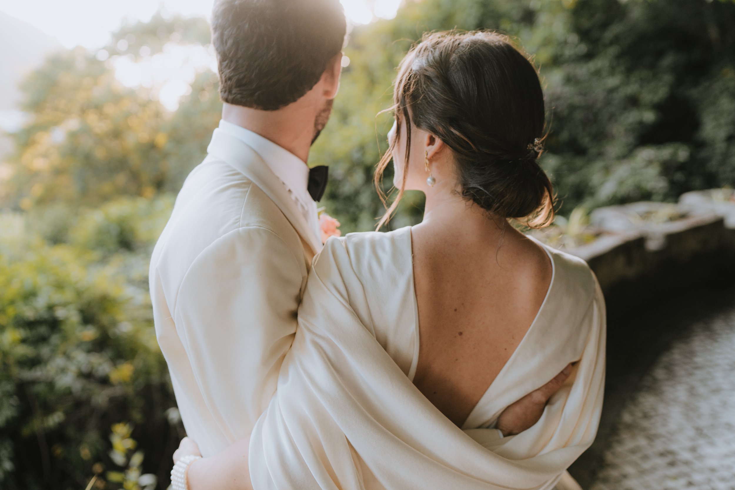 A couple in wedding attire embracing outdoors near a river, with lush greenery and sunlight in the background.
