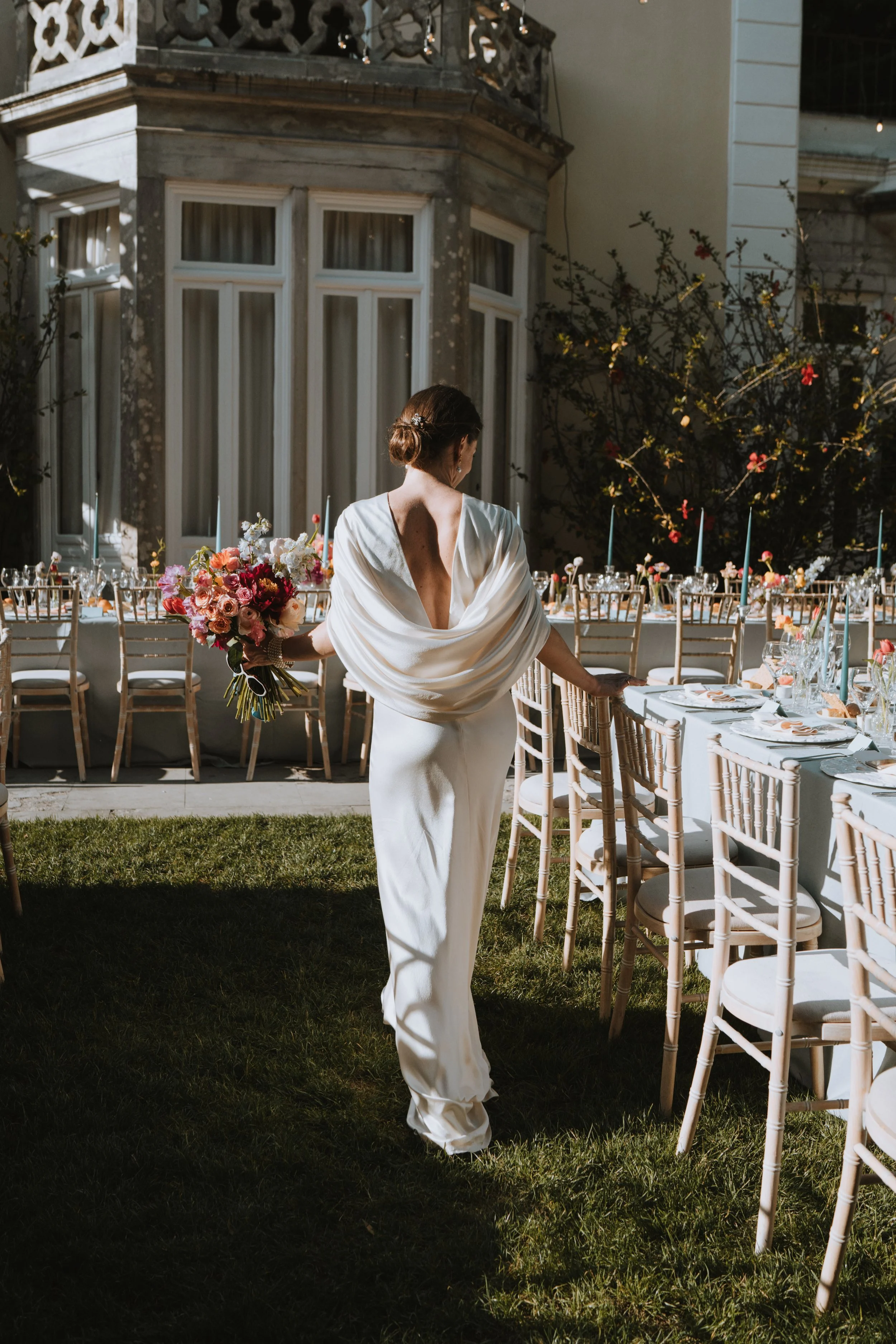 A woman in a white gown holding a bouquet of colorful flowers at an outdoor wedding reception with decorated tables and chairs.