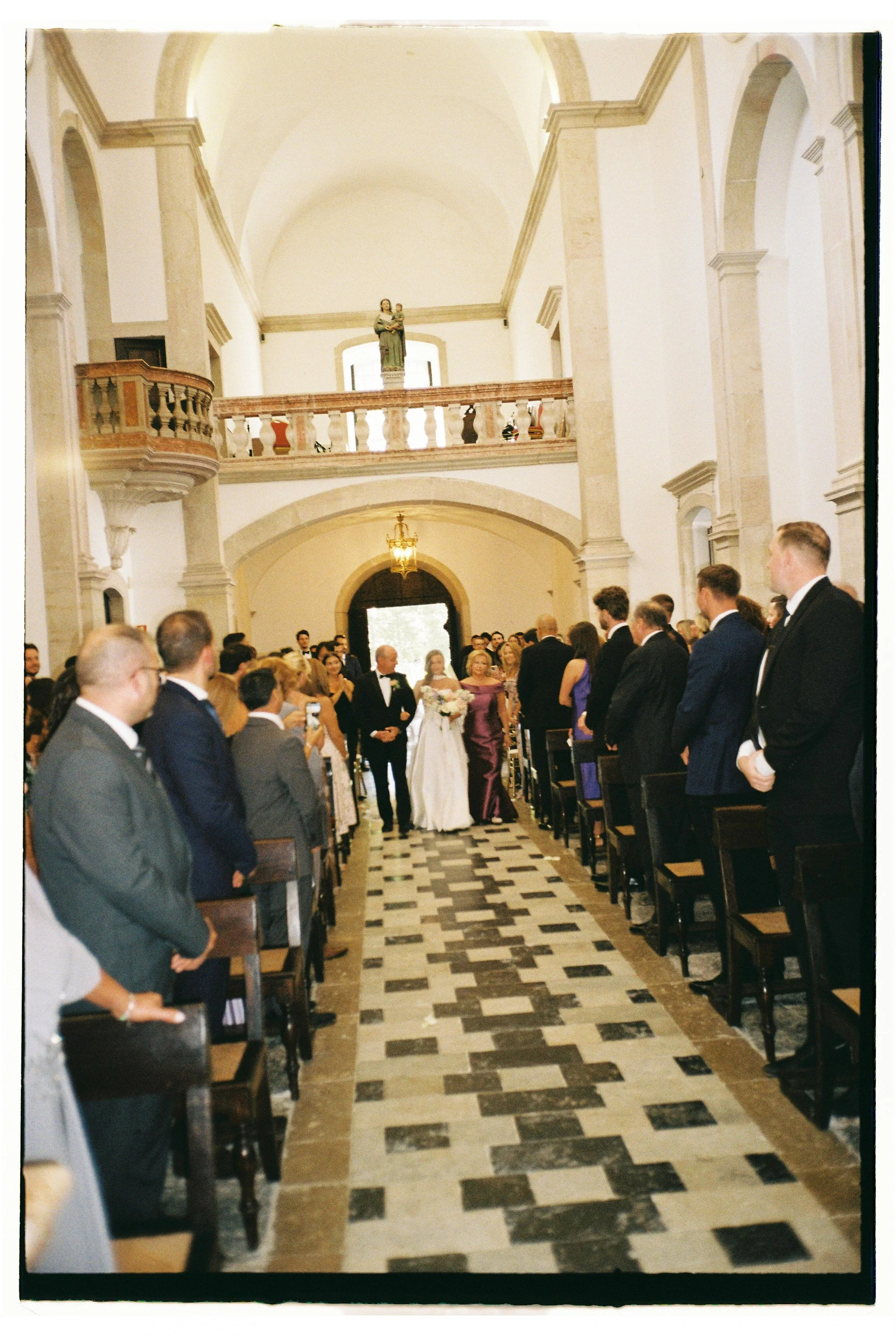 A wedding ceremony is taking place inside a church with high vaulted ceilings. The bride and groom are walking down the aisle, surrounded by family and friends. The church has a second-floor balcony with a statue on display, and the floor features a 