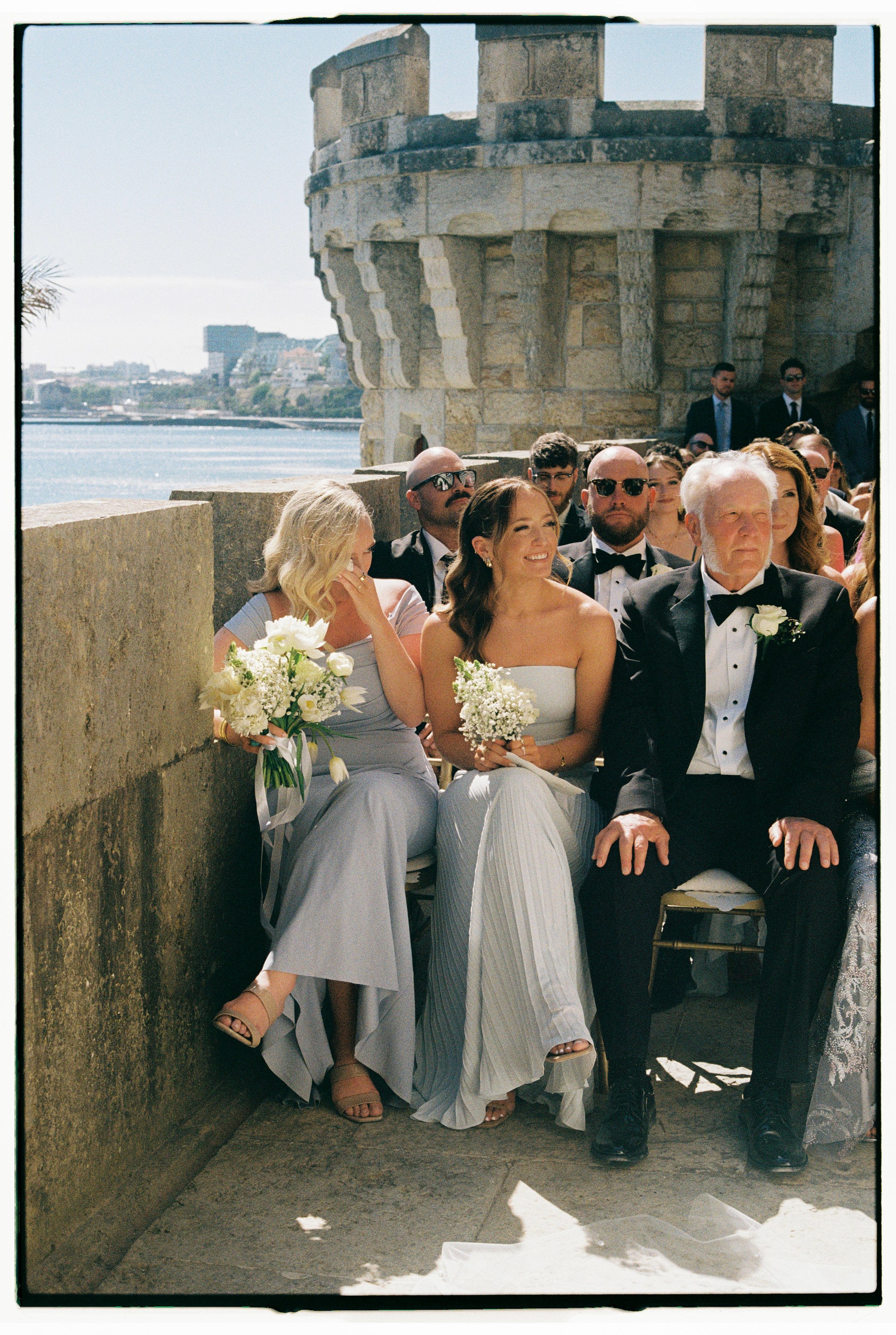 A wedding ceremony taking place outdoors by a river, with guests dressed formally. The bride and groom sit together, with the bride holding a bouquet of white flowers, and an older man in a tuxedo and white boutonniere sitting beside her. Other guest
