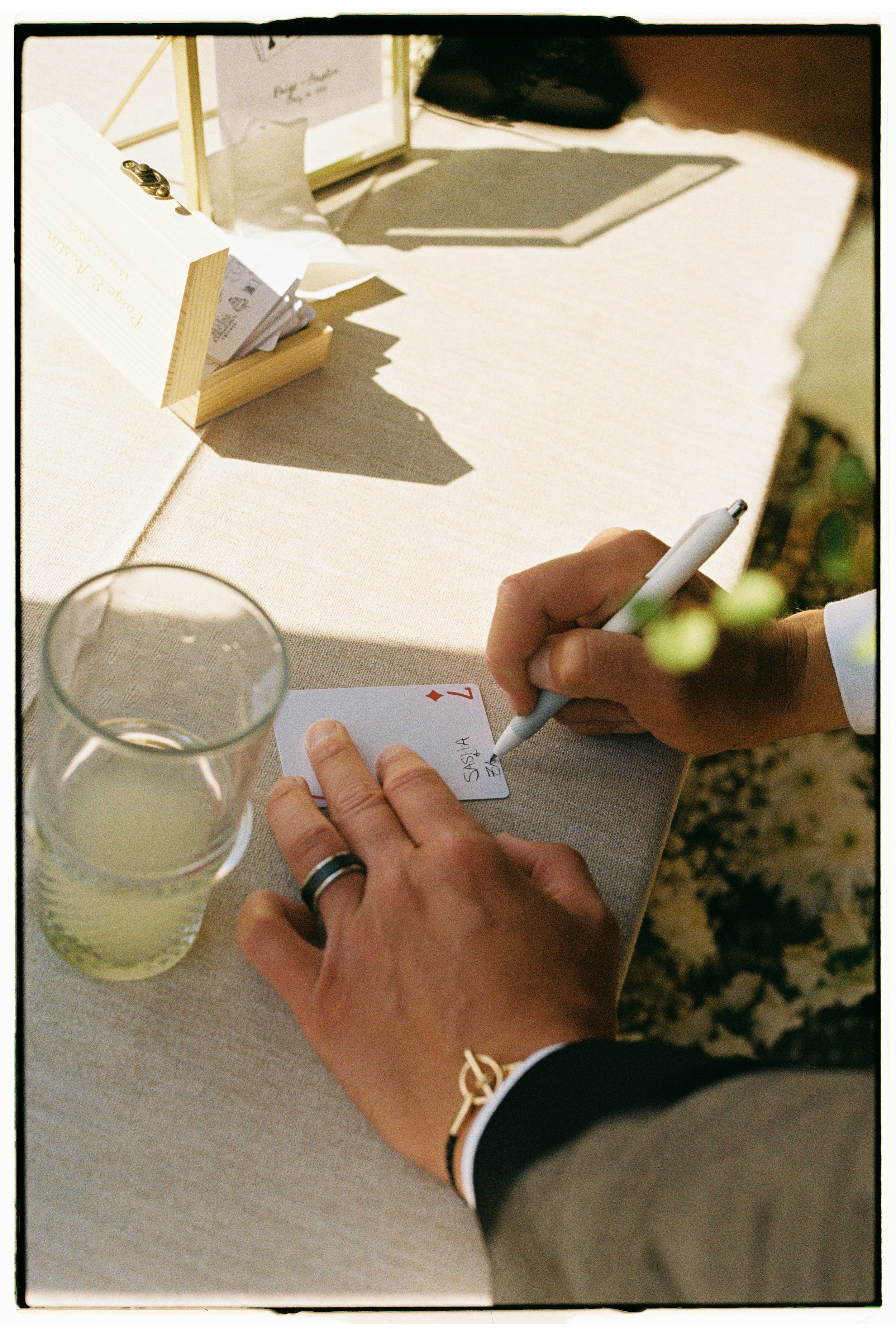 Person signing a playing card with a white pen at a table, with a glass of beverage and various papers and items nearby.