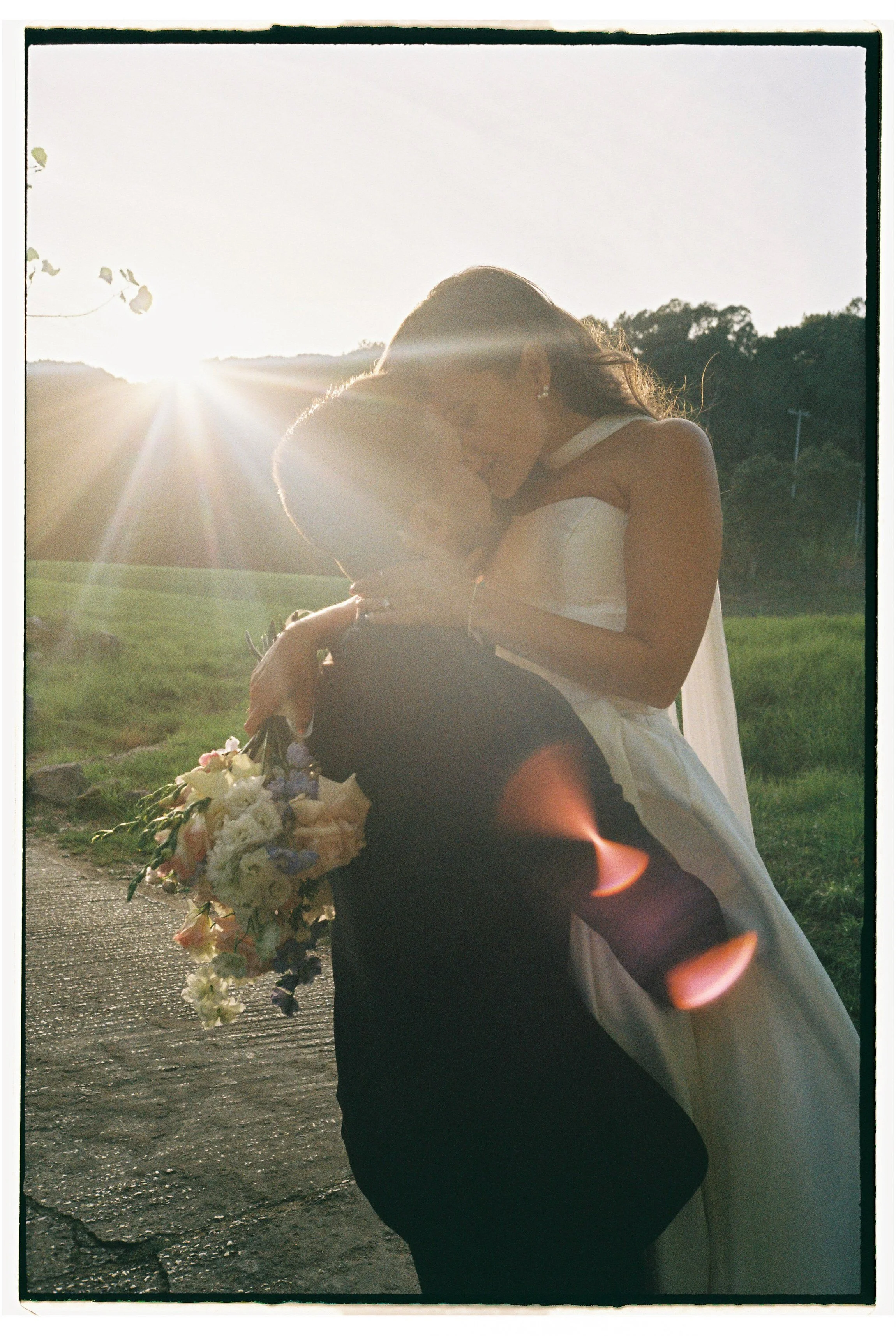 A bride and groom share a romantic kiss outdoors during sunset, with the bride holding a bouquet of flowers and the sun creating a lens flare.