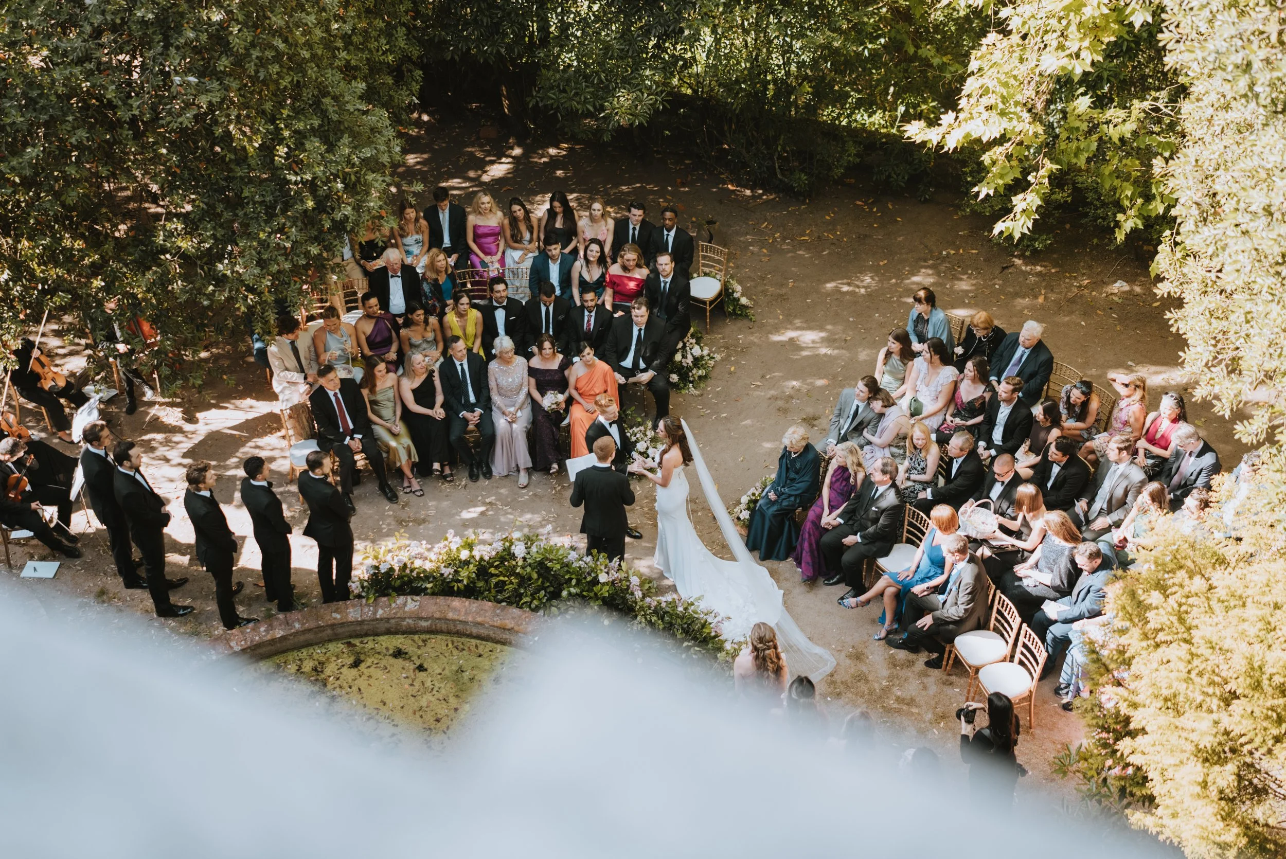 An outdoor wedding ceremony with guests seated on either side of the bride and groom, surrounded by trees and greenery.