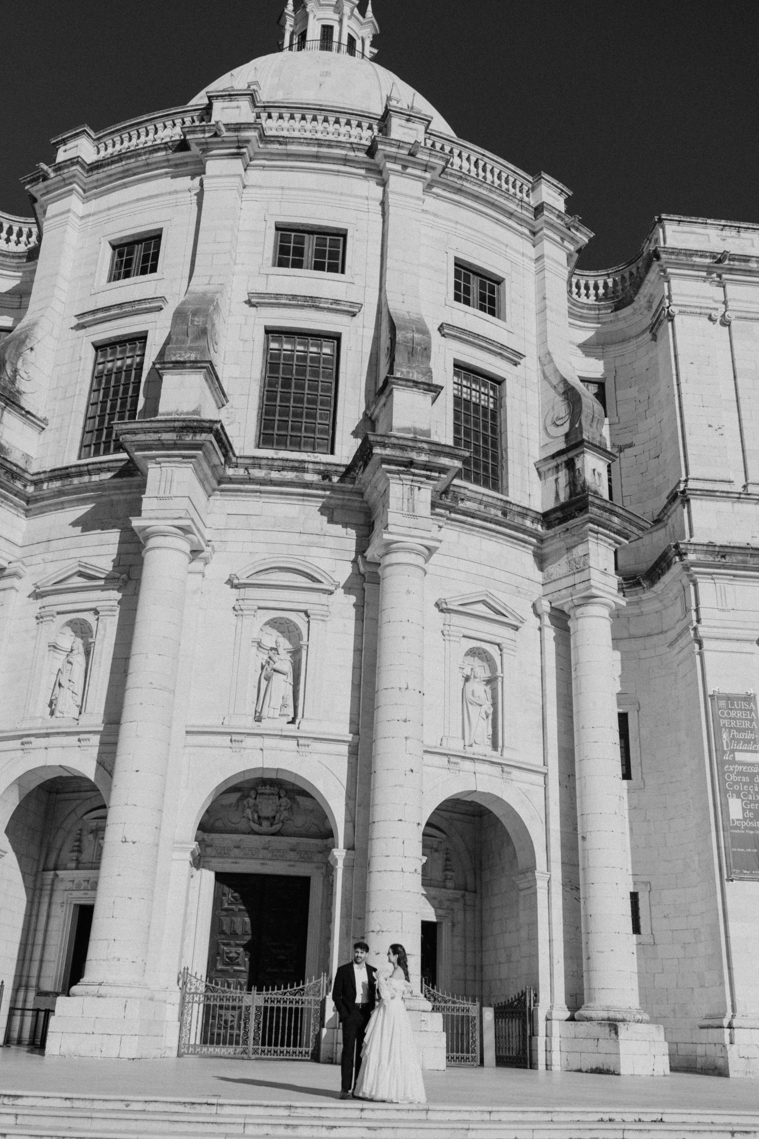 A black and white photo of a large historic church or cathedral with a rounded dome, tall columns, and statues on the facade. In front, a couple dressed in formal wedding attire stands on steps.