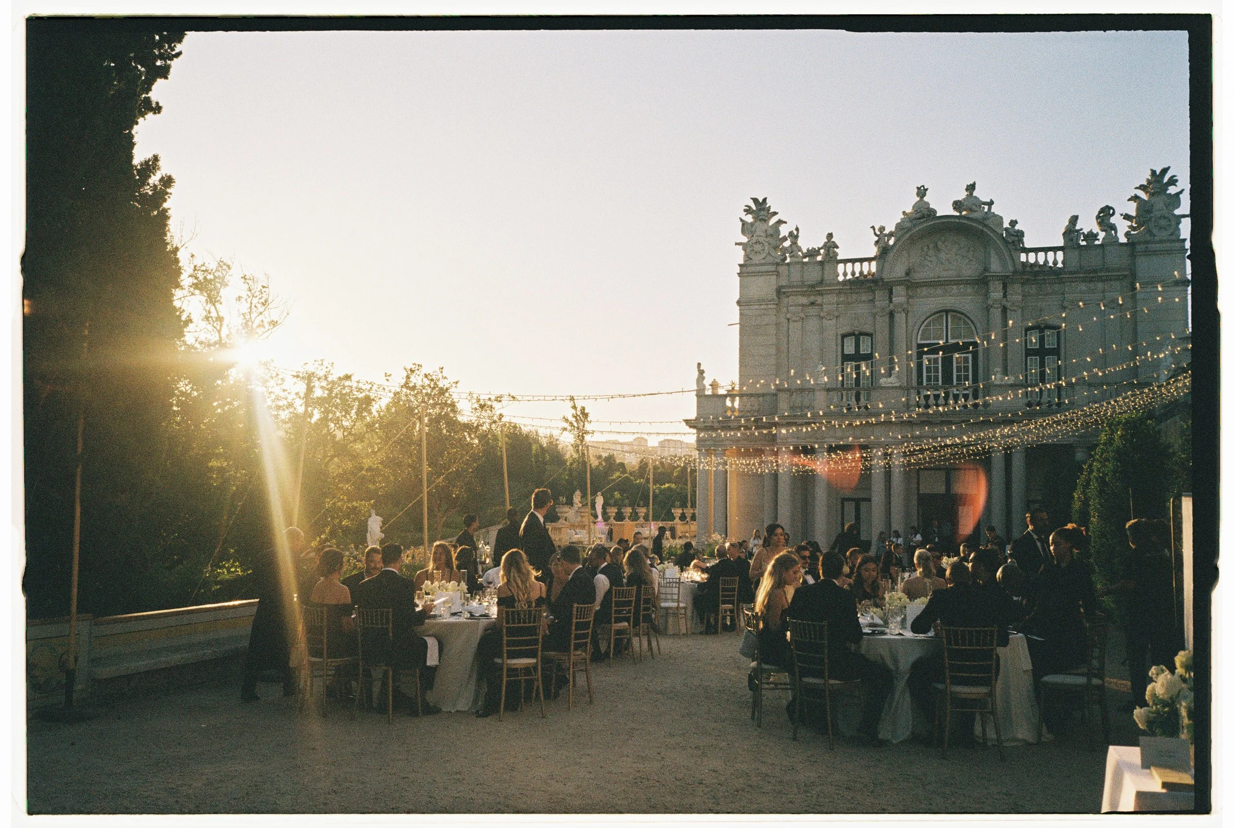 An outdoor evening event with guests seated at round tables under string lights in front of a historic building with ornate architecture, surrounded by greenery.