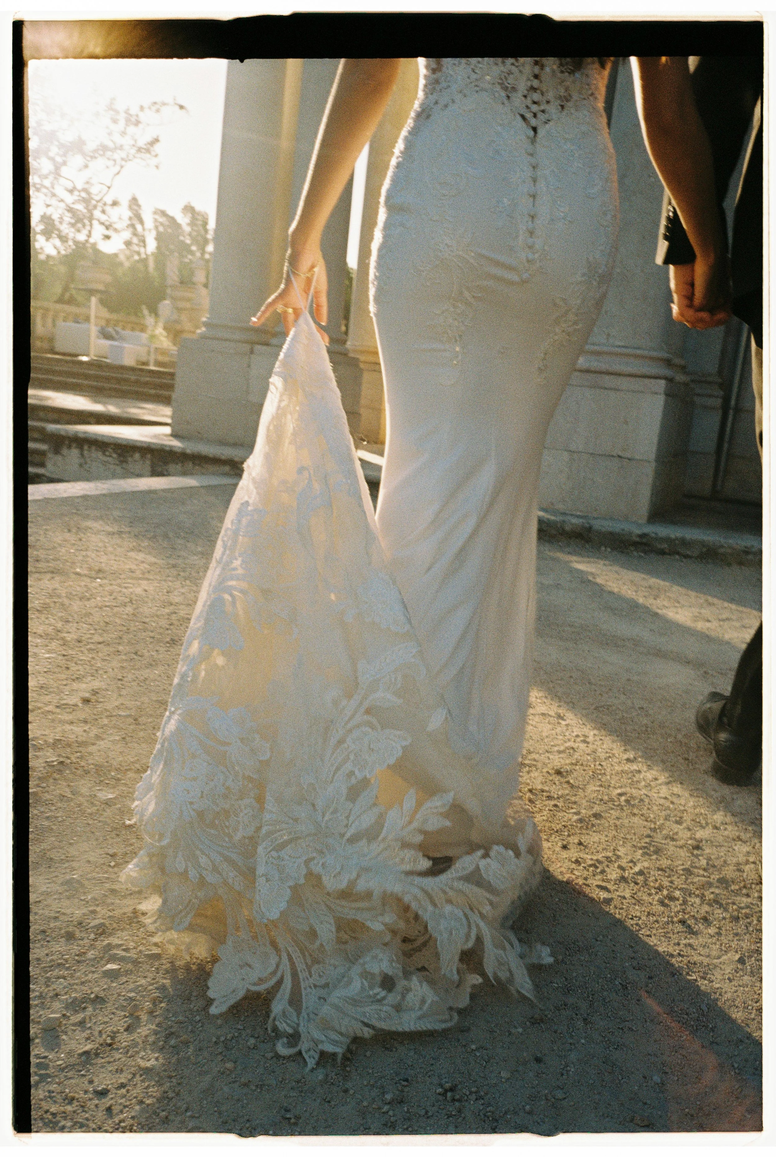 Bridal couple holding hands, with the bride lifting her lace wedding train, with the sun setting in the background.
