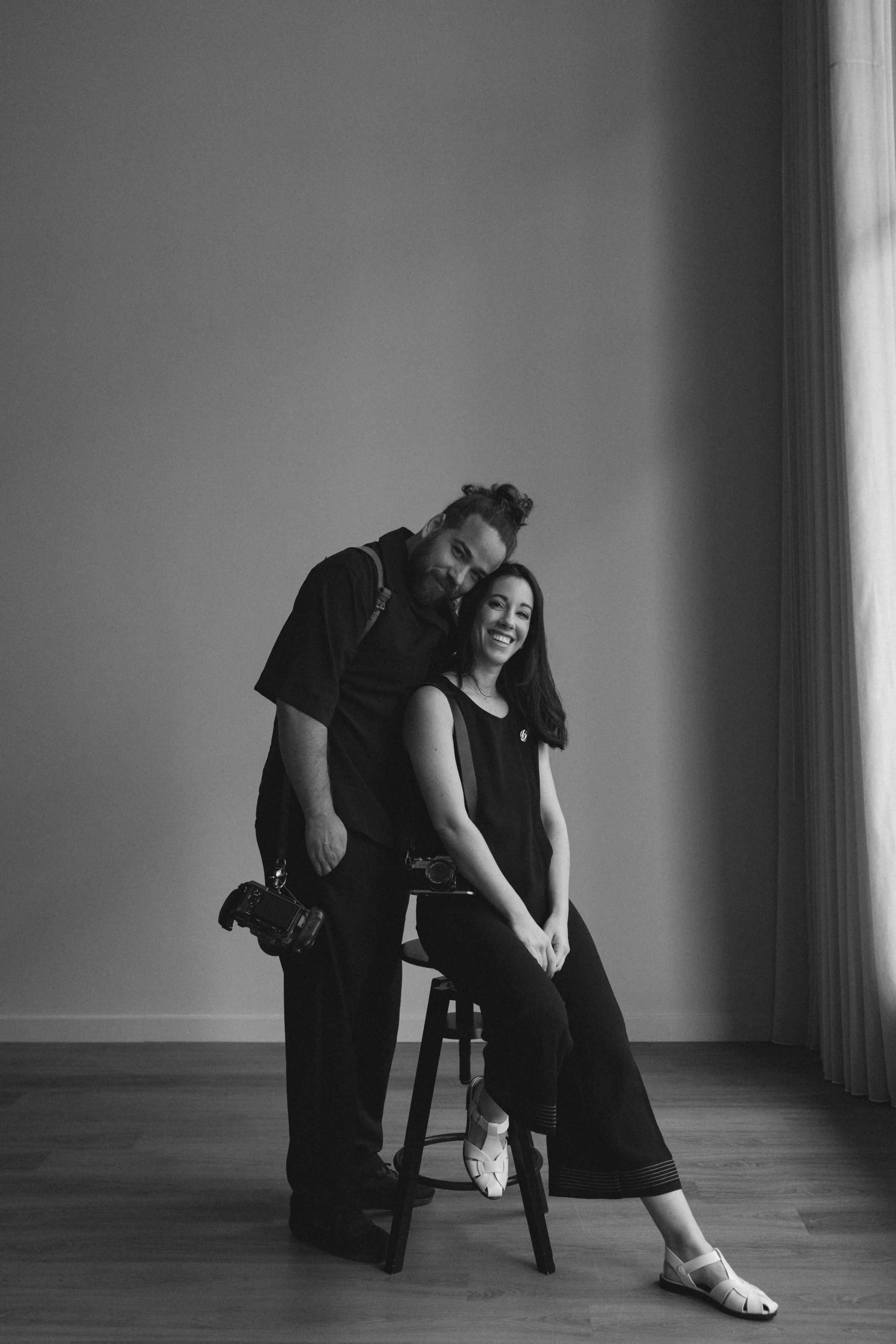 A black and white photo of a man and woman posing indoors against a plain wall and curtain. The woman is seated on a stool, smiling, while the man stands beside her with his head gently resting on her head. Both are casually dressed, and the woman is holding a camera.