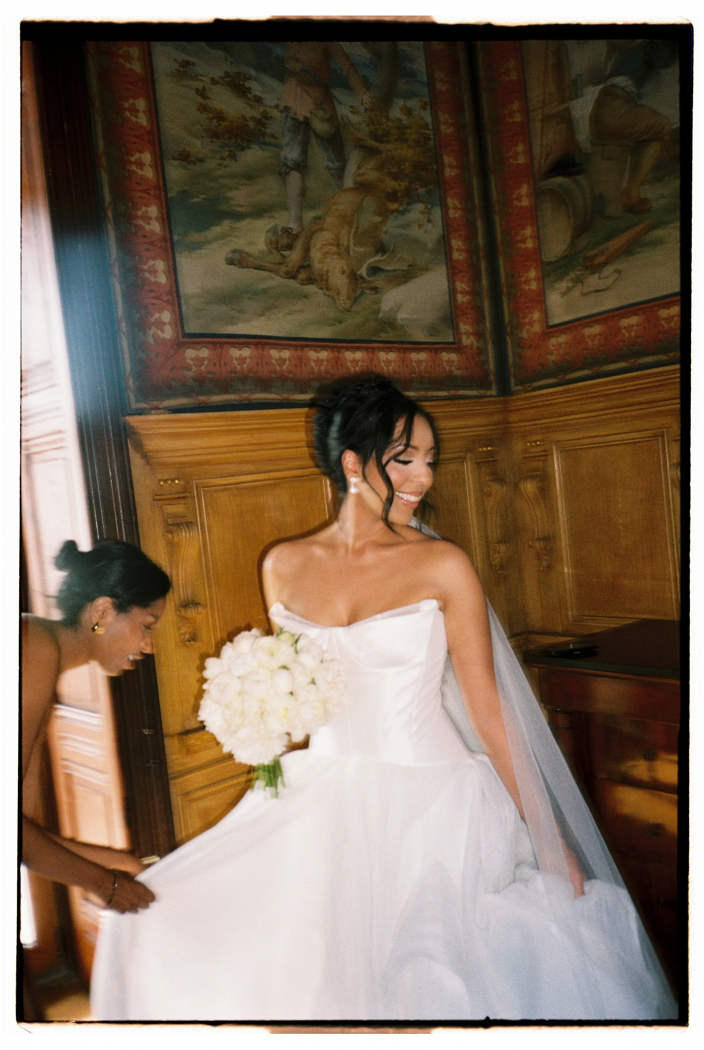 A bride in a white wedding gown holding a bouquet of white flowers, smiling, with another woman assisting her in a room with wooden paneling and a ceiling mural of a historical scene.