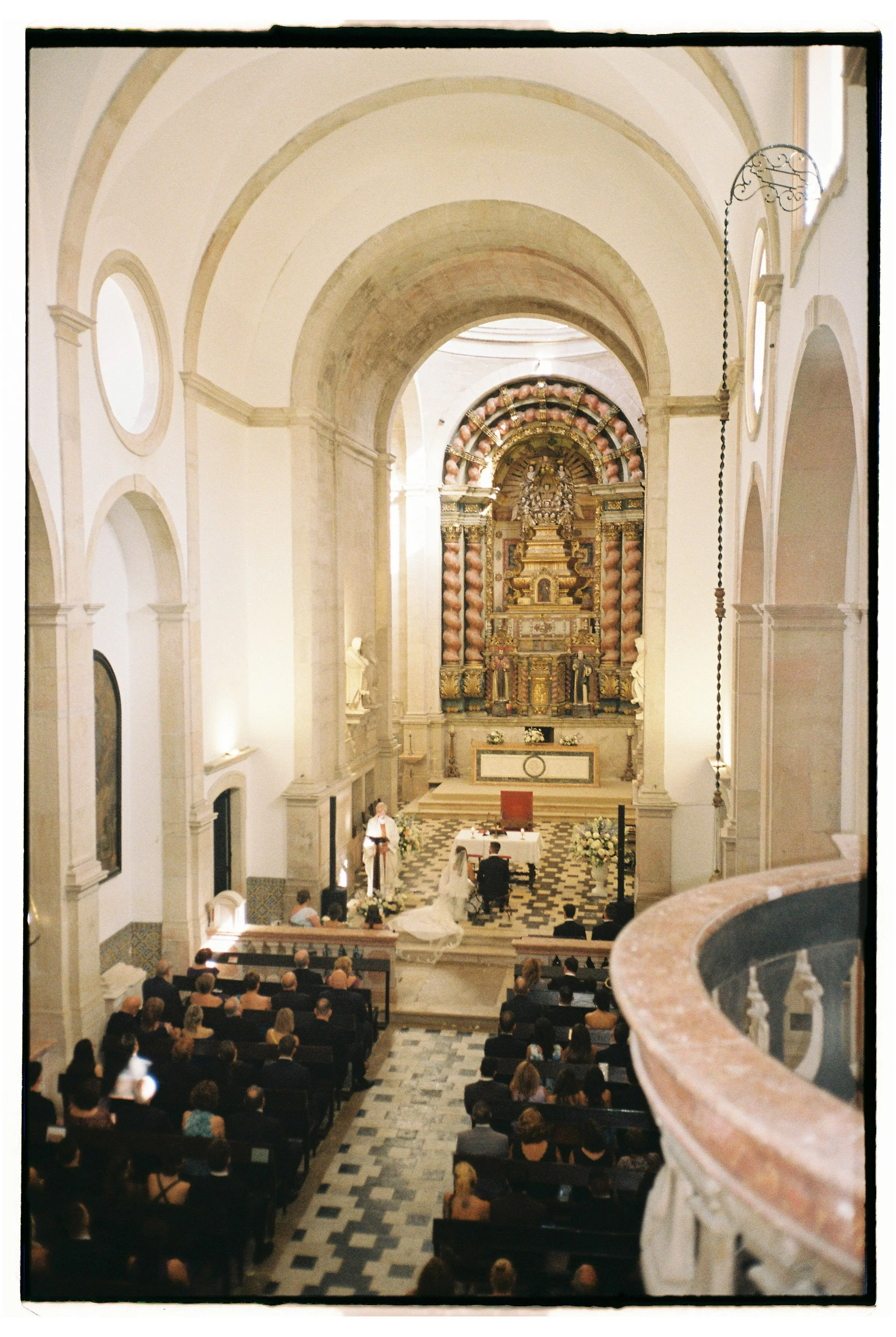 Interior of a church with a wedding ceremony in progress, featuring a bride and groom kneeling at the altar, guests seated in pews, and ornate decorations at the altar.