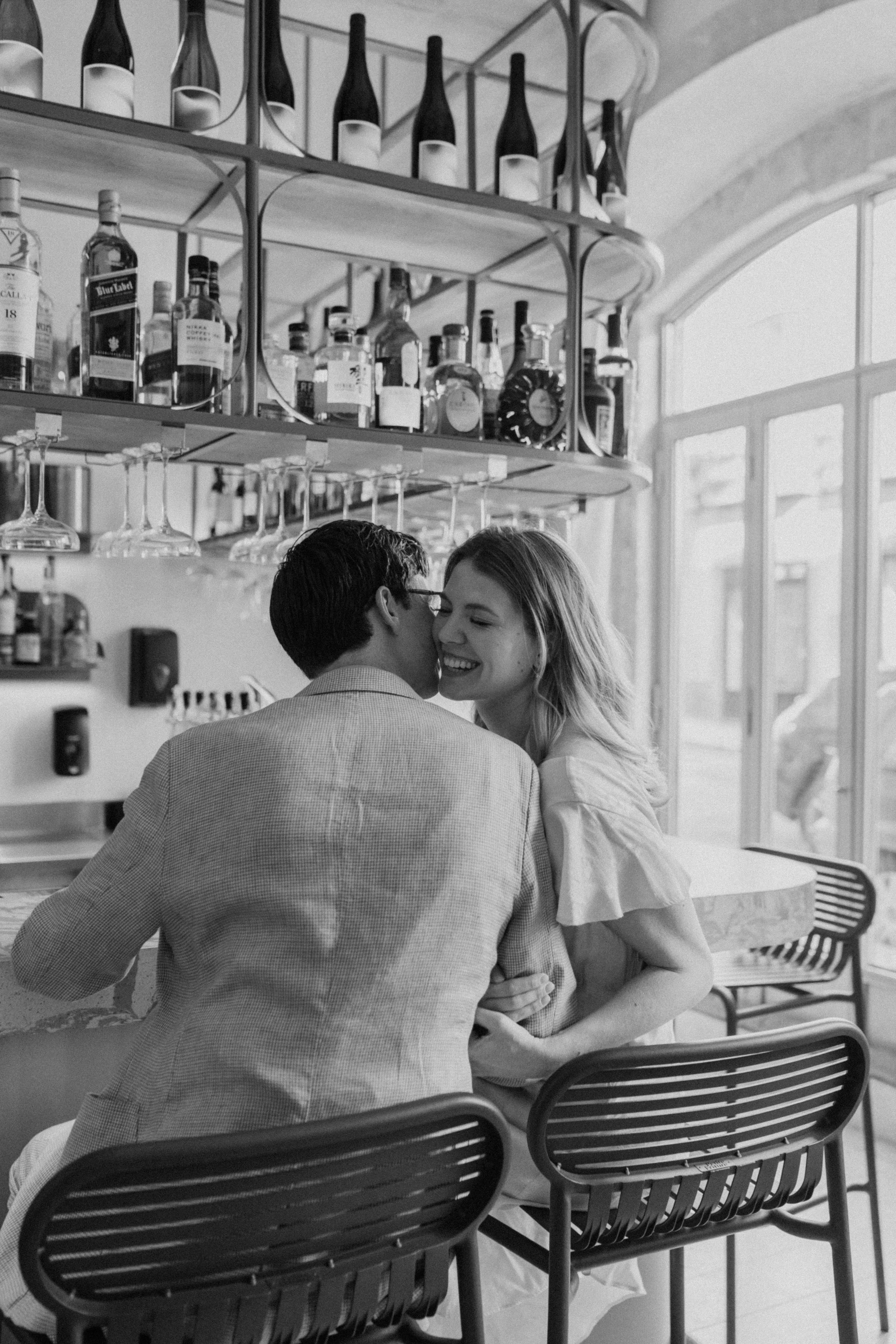 A black and white photo of a couple sitting closely at a bar, smiling and sharing an intimate moment, with shelves of liquor bottles and wine glasses behind them, and large windows letting in natural light.